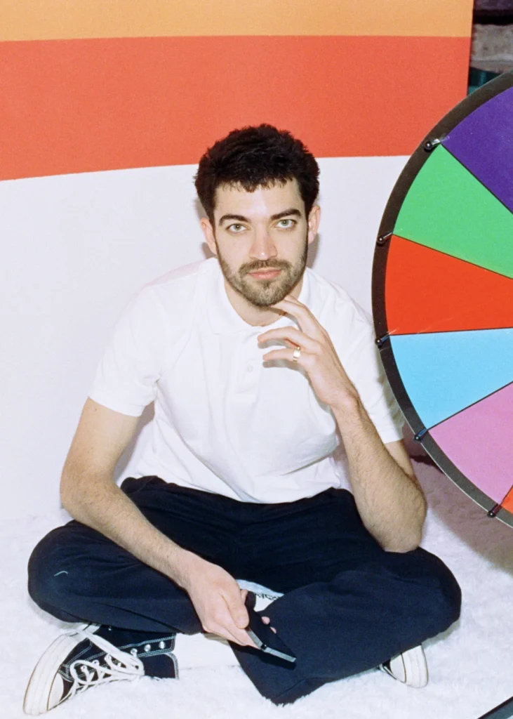 Adam Faze sitting on a white floor with a colorful prize wheel and a striped orange and yellow backdrop.