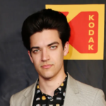 A young man in a gray blazer posing in front of a Kodak logo, known for his career and net worth.