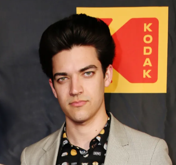 A young man in a gray blazer posing in front of a Kodak logo, known for his career and net worth.