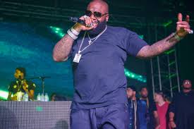 Rick Ross performing live on stage, holding a microphone while wearing a blue outfit and chain necklace.